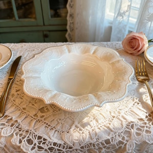 May include: A white ceramic serving bowl with a scalloped edge and embossed floral design. The bowl is on a lace tablecloth, with a gold fork and knife visible. A pink rose is in the background.