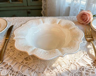 Könnte beinhalten: Eine weiße Keramik-Servierschüssel mit gewelltem Rand und geprägtem Blumendesign. Die Schüssel steht auf einer Spitzendecke, mit einer goldenen Gabel und einem Messer. Im Hintergrund ist eine rosa Rose zu sehen.