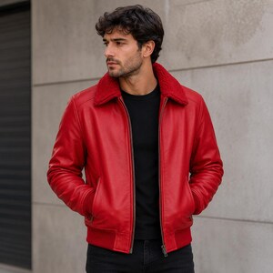 May include: A bright red leather bomber jacket with a red shearling collar. The jacket has a zip-up front, two front pockets, and ribbed cuffs and hem. The model is wearing a black top and black trousers.