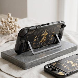 Może przedstawiać: Czarna obudowa konsoli Nintendo Switch ze złotym wzorem przedstawiającym miecz i inne symbole. Obudowa jest podparta na szarej podstawce. Poniżej znajduje się druga obudowa. Projekt przypomina grę wideo.