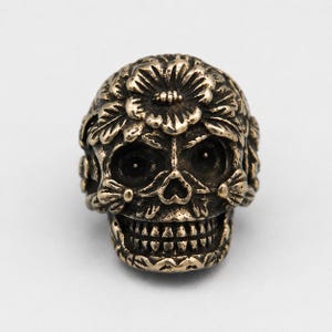 May include: A metallic bronze-toned skull bead featuring intricate relief carvings of flowers and leaves across the cranium and cheekbones. The skull has deep, dark eye sockets, a small nasal cavity, and a detailed row of teeth in the lower jaw.