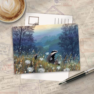 Puede incluir: Una postal con una ilustración de tejones en un campo. El fondo muestra un paisaje brumoso con árboles. También se ven una taza de café y un bolígrafo.