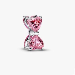 Könnte beinhalten: Ein silberner Charm mit zwei facettierten, herzförmigen rosa Steinen, die vertikal übereinander angeordnet sind. Die Steine werden von silbernen Krappen gehalten und durch ein mittleres Silberband verbunden, was eine symmetrische Sanduhrform ergibt.