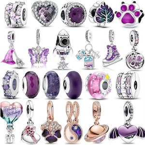 Puede incluir: Una colección de veinticuatro dijes de metal plateado con diversas formas, incluyendo corazones, un cohete, una huella, una hoja, una mariposa, un sombrero de bruja, patines, un símbolo yin-yang y un planeta, decorados con esmalte morado y cristales.