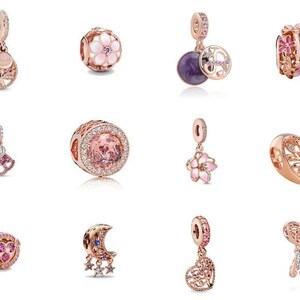 Könnte beinhalten: Eine Sammlung von zwölf Anhängern aus roségoldfarbenem Metall mit verschiedenen Motiven, darunter ein Lebensbaum, florale Muster mit rosa Emaille, eine violette runde Scheibe, ein herzförmiger Anhänger, eine Mondsichel mit Sternen und ein Traumfänger.
