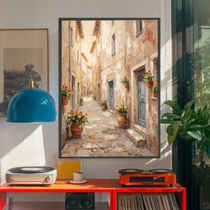 May include: Oil painting of a narrow European street scene. The artwork depicts buildings with textured walls, blue doors, and flower pots. The cobblestone street leads to a distant figure. The painting is framed in black and hangs on a white wall.