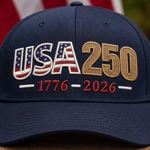 以下が含まれることがあります： 前面に刺繍が施されたネイビーブルーの野球帽。USAの文字は赤、白、青の星条旗柄で埋め尽くされています。その横には金色の刺繍で250という数字があり、下部には赤色で1776と2026という年号が刺繍されています。