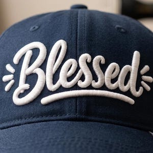 Puede incluir: Gorra de béisbol azul marino con la palabra "Blessed" bordada en blanco en la parte delantera. La palabra tiene un diseño estilizado con pequeñas explosiones de luz a cada lado. La gorra tiene una visera curva y una pequeña etiqueta con la bandera americana.
