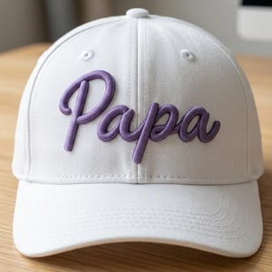 May include: A white baseball cap featuring the word Papa embroidered in raised purple script lettering across the front panels. The cap has a structured crown, a curved brim, and stitched eyelets for ventilation on the top panels.