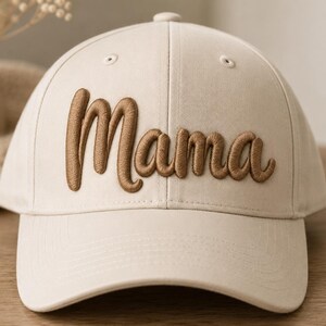 May include: A beige baseball cap featuring the word Mama embroidered in raised, brown, cursive lettering across the front panel. The cap has a structured crown, a curved brim, and stitched eyelets on the top panels for ventilation.