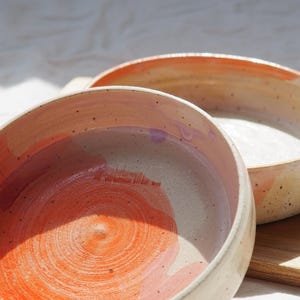 Könnte beinhalten: Zwei runde Keramikschalen mit gesprenkelter cremefarbener Basis, die innen mit abstrakten orangefarbenen und hellvioletten Pinselstrichen versehen sind. Die vordere Schale zeigt ein schwaches, eingeprägtes Logo in der Mitte.