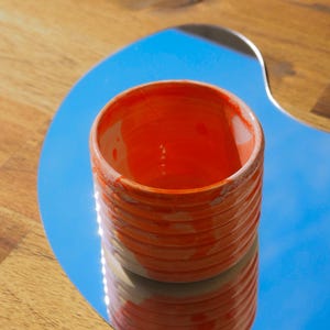 Könnte beinhalten: Ein zylindrischer Keramikbecher mit einer leuchtend orangefarbenen Glasur, glänzender Oberfläche und horizontaler Rippenstruktur an der Außenseite. Der Becher hat eine weite, kreisförmige Öffnung und steht auf einer reflektierenden Oberfläche.