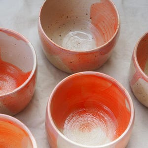 Könnte beinhalten: Eine Sammlung von fünf Keramikschalen mit einer gesprenkelten cremeweißen Basis und handgemalten, leuchtend orangefarbenen Pinselstrichen. Jede Schale hat eine abgerundete Form und eine glatte, glänzende Oberfläche mit sichtbarer Textur und Variationen.