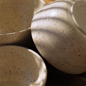 Könnte beinhalten: Drei Keramikschalen mit einer cremefarbenen Glasur und braunen Sprenkeln. Die Schalen haben eine abgerundete Form und eine glatte, glänzende Oberfläche. Das Licht wirft lineare Schatten auf die Oberfläche der Schalen und betont ihre Wölbung.