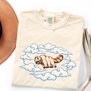 Könnte beinhalten: Ein cremefarbenes Kurzarm-T-Shirt mit einer Grafik eines pelzigen, sechsbeinigen Wesens mit braun-weißem Fell, das inmitten stilisierter, blau umrandeter Wolken fliegt. Das innere Nackenetikett zeigt den Text Comfort Colors.