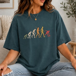 May include: A dark teal short-sleeve t-shirt featuring a graphic print of the human evolution sequence, progressing from a primate to a human figure bowling with a red ball. The design uses muted green, beige, orange, yellow, and red colors.