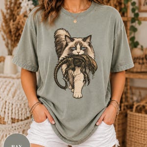 Puede incluir: Una camiseta de manga corta color verde salvia con un estampado gráfico de un gato peludo que lleva una criatura biomecánica oscura en la boca. La camiseta tiene cuello redondo y un corte holgado, con la etiqueta de color BAY en un círculo.