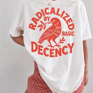Könnte beinhalten: Ein weißes T-Shirt mit einer roten Grafik eines Vogels, der einen Zweig hält. Der Text RADICALIZED BY BASIC DECENCY ist in Rot um den Vogel herum gedruckt. Das T-Shirt wird mit einer rot-weiß karierten Shorts im Vichy-Muster kombiniert.