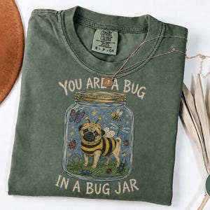 Op de afbeelding: Een mosgroen t-shirt met korte mouwen en een afbeelding van een mopshond in een bijenkostuum in een glazen pot. De tekst luidt YOU ARE A BUG IN A BUG JAR. Het ontwerp bevat ook een vlinder, een bij, een libel en een lieveheersbeestje op de stof.