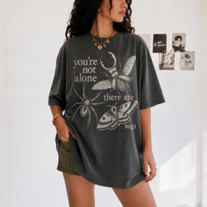 Pode incluir: Uma t-shirt cinzento-carvão de corte oversize com uma impressão gráfica de um escaravelho, uma aranha e uma traça. O texto diz, you're not alone there are bugs. A t-shirt tem mangas curtas e gola redonda, combinada com calções de veludo cotelê verde-azeitona.