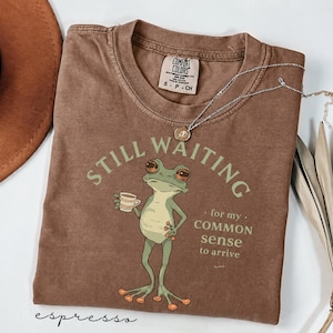 May include: A brown short-sleeve t-shirt featuring a graphic of a green frog holding a small cup. The text reads STILL WAITING for my COMMON SENSE to arrive. The shirt has a Comfort Colors brand label at the neck and a crew neckline design.