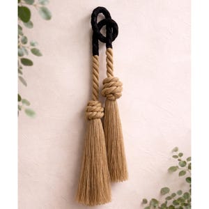 May include: Decorative wall hanging featuring two long tassels made of natural-colored rope. The top section is black, transitioning to a twisted rope design. Each tassel has a large knot and a long, fringed end, creating a textured, rustic aesthetic.