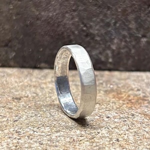Könnte beinhalten: Ein silberner Ring mit facettiertem Design. Der Ring besteht aus einem glänzenden, metallischen Material und hat einen schlichten, eleganten Stil. Der Ring wird auf einer strukturierten, neutral getönten Oberfläche gezeigt.