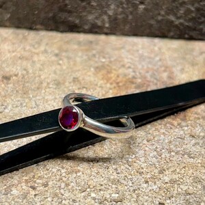 Könnte beinhalten: Ein silberner Ring mit einem runden, tiefroten Edelstein. Der Ring liegt auf einer schwarzen Oberfläche vor einem strukturierten, hellbraunen Hintergrund. Der Edelstein ist der Blickfang und zeigt seine satte Farbe und seinen Glanz.