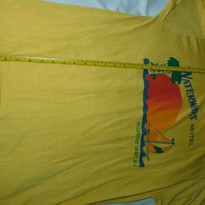 May include: Yellow t-shirt with a graphic of a sailboat, sun, and the text "Waterways Motel Wildwood Crest N.J." A measuring tape is laid across the shirt, indicating its length.