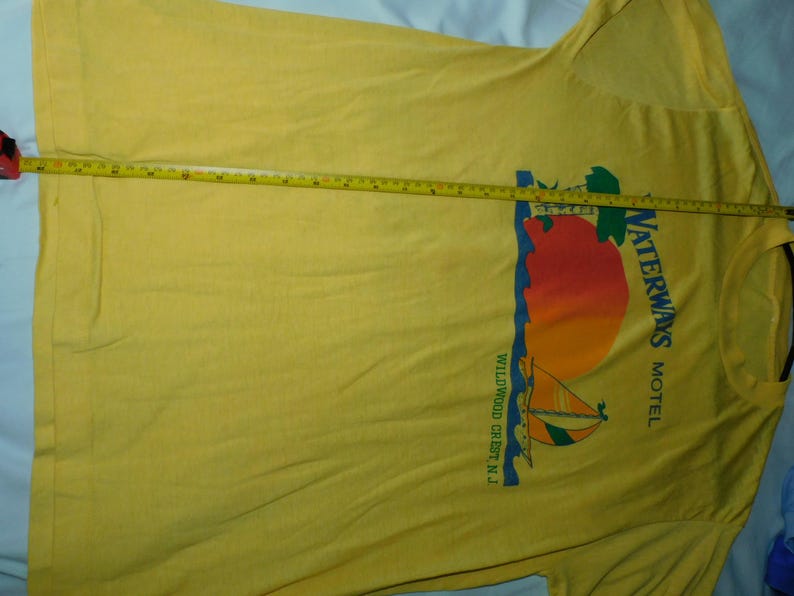 May include: Yellow t-shirt with a graphic of a sailboat, sun, and the text "Waterways Motel Wildwood Crest N.J." A measuring tape is laid across the shirt, indicating its length.
