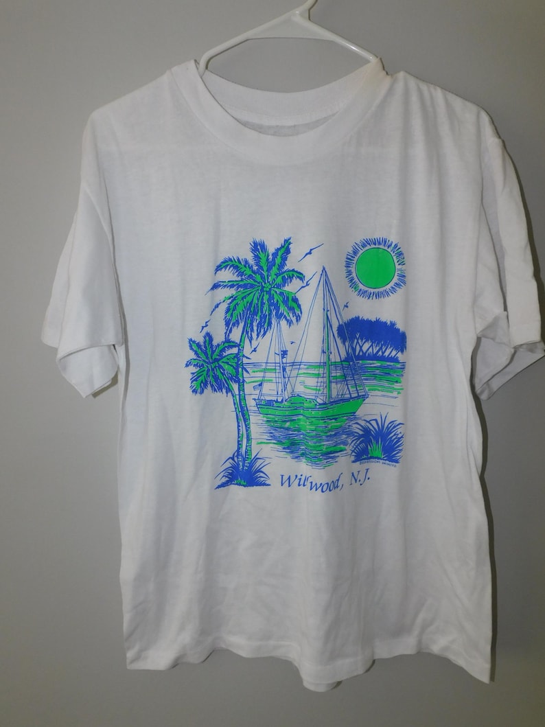 May include: White t-shirt featuring a graphic of a sailboat, palm trees, and a sun in shades of blue and green. The text "Wildwood, N.J." is printed below the image. The shirt is hanging on a white hanger.