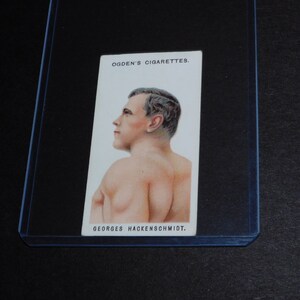 May include: A vintage Ogden's Cigarettes trading card featuring a portrait of Georges Hackenschmidt. The card shows a man from the chest up, facing left, with a detailed illustration. The card is in a protective sleeve.