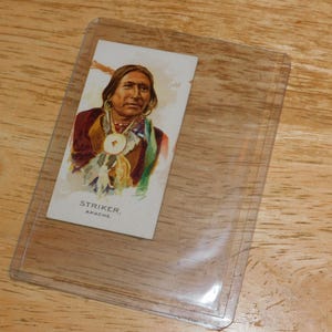 May include: A vintage trading card featuring a portrait of Striker, Apache. The card is in a protective sleeve. The portrait shows a man with long dark hair, wearing a necklace and a colorful garment. The card is on a wooden surface.