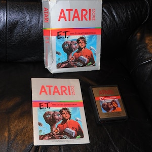 May include: An Atari 2600 E.T. The Extra-Terrestrial video game set including a white cardboard box, a paper instruction manual, and a black plastic game cartridge. Each item features the Atari logo and artwork of E.T. and a young boy.