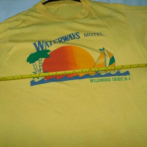 May include: Yellow t-shirt with a graphic featuring a sunset, sailboat, and palm trees. The text "WATERWAYS MOTEL WILDWOOD CREST N.J." is printed above and below the graphic. A yellow tape measure is laid across the shirt.