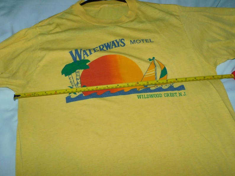 May include: Yellow t-shirt with a graphic featuring a sunset, sailboat, and palm trees. The text "WATERWAYS MOTEL WILDWOOD CREST N.J." is printed above and below the graphic. A yellow tape measure is laid across the shirt.