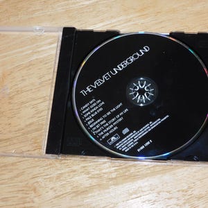 May include: A compact disc (CD) of The Velvet Underground album, in a clear plastic jewel case. The black CD has the band's name and song titles printed on it. The case is open, resting on a wooden surface.