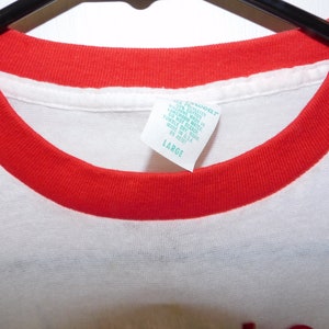 May include: A white t-shirt with a red crew neck collar. The tag reads "LARGE". The shirt is made of a soft material. The shirt is hanging on a black hanger.