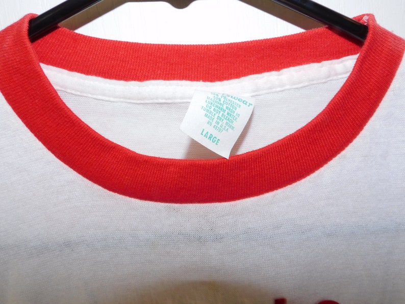 May include: A white t-shirt with a red crew neck collar. The tag reads "LARGE". The shirt is made of a soft material. The shirt is hanging on a black hanger.