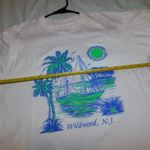 May include: White t-shirt featuring a sailboat scene with palm trees, sun, and the text "Wildwood, N.J." in blue and green. The shirt is on a hanger, and a measuring tape is across the shirt.