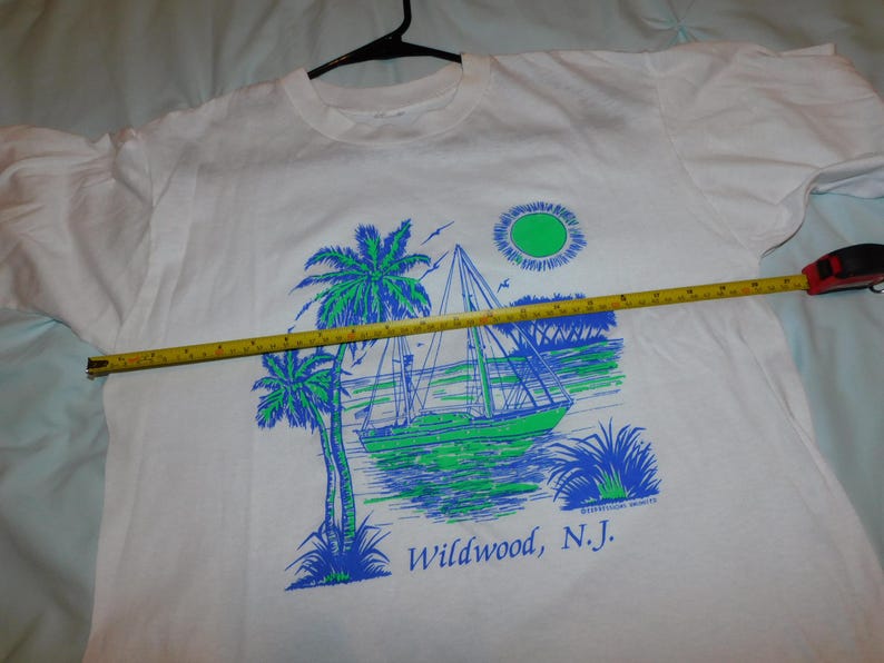 May include: White t-shirt featuring a sailboat scene with palm trees, sun, and the text "Wildwood, N.J." in blue and green. The shirt is on a hanger, and a measuring tape is across the shirt.