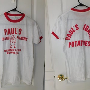 May include: White t-shirt with red trim featuring the text "Paul's Idaho Potatoes" in red, along with a cartoon potato character. The shirt also includes the text "Boardwalk & Cedar Wildwood, N.J."