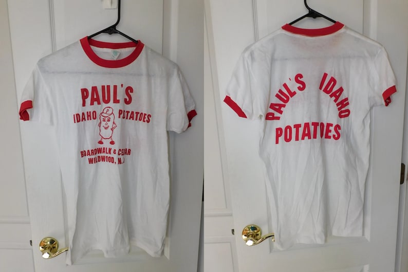 May include: White t-shirt with red trim featuring the text "Paul's Idaho Potatoes" in red, along with a cartoon potato character. The shirt also includes the text "Boardwalk & Cedar Wildwood, N.J."
