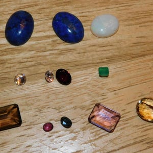 May include: A collection of loose gemstones in various shapes and colors, including two large oval deep blue stones, a smooth white opal, a small square green stone, several round faceted gems, and three larger rectangular and oval faceted stones in brown and pink.