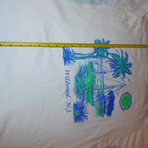 May include: White t-shirt featuring a blue and green graphic of palm trees, sailboats, and a sun. The text "Wildwood, N.J." is printed vertically on the right side. A yellow tape measure is laid across the shirt.