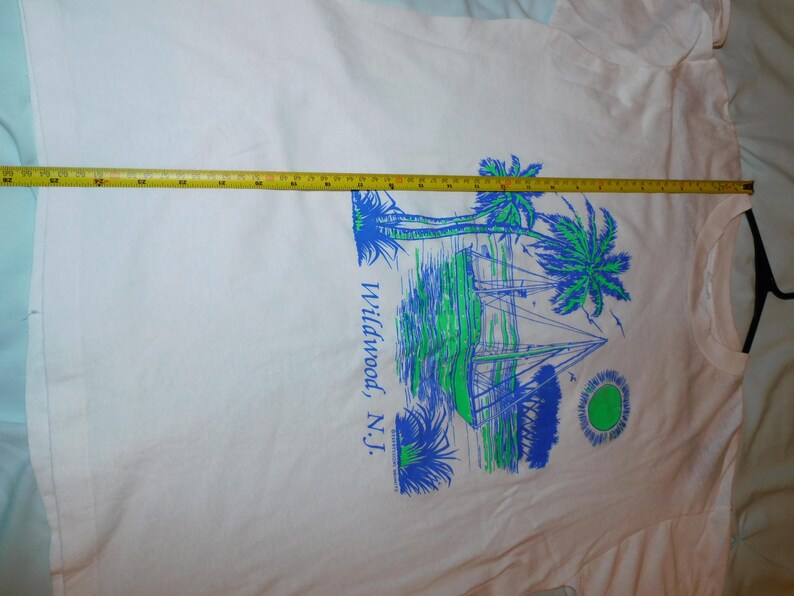May include: White t-shirt featuring a blue and green graphic of palm trees, sailboats, and a sun. The text "Wildwood, N.J." is printed vertically on the right side. A yellow tape measure is laid across the shirt.
