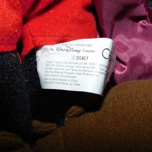 May include: Close-up of a plush toy with red, black, and brown fabric. A white tag with text from The Walt Disney Company is visible. The tag includes the Disney logo and manufacturing information.
