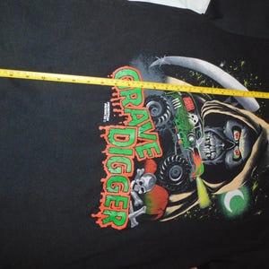 Może przedstawiać: Czarna koszulka z grafiką monster trucka Grave Digger i ponurego żniwiarza z kosą. Napis "Grave Digger" jest w kolorze zielonym i pomarańczowym. Koszulka ma około 76 cm szerokości.