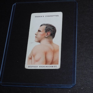 May include: A vintage Ogden's Cigarettes trading card featuring Georges Hackenschmidt. The card shows a side profile of a man with short hair and a muscular physique. The card is in a protective sleeve.