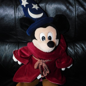 May include: A plush Mickey Mouse dressed as a sorcerer, wearing a dark blue hat with white stars and a crescent moon. He has a burgundy velvet robe and brown shoes. The toy is on a black leather surface.